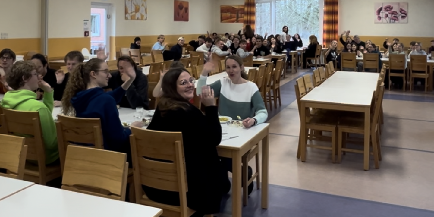 Startschuss für die Schultheaterwoche 2025!  – Auf dem Bild ist der Essensraum der Jugendherberge Flensburg zusehen. In dem Raum stehen mehrere Tische und Stühle an denen Schüler und Schülerinnen sitzen. Diese gucken alle lächelnd und winkend in Richtung der Kamera. 